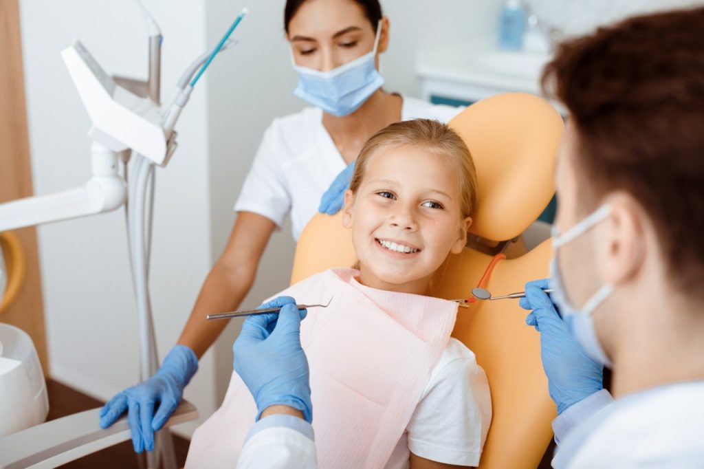 Cheerful little kid in dental chair looks at millennial male dentist in white coat